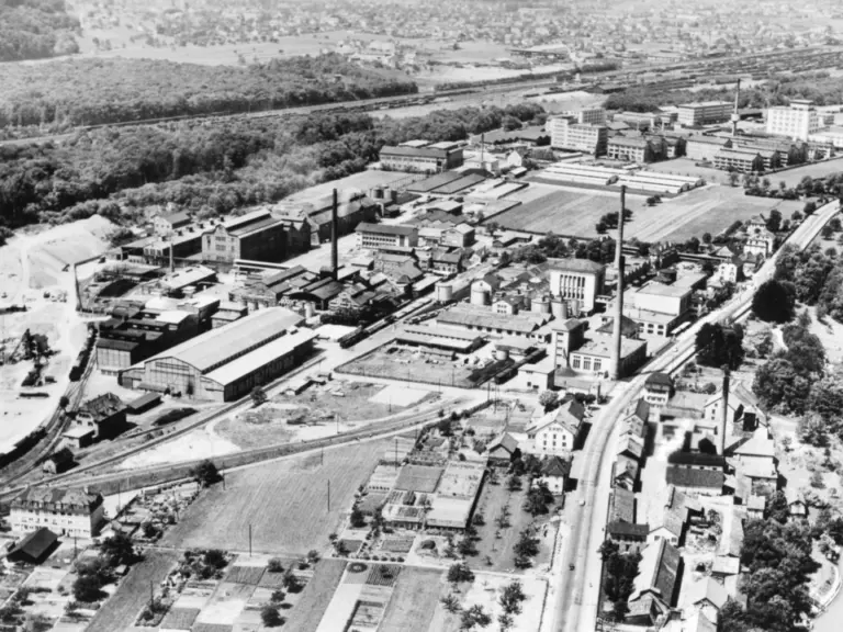 Schwarzweisse Luftaufnahme eines weitläufigen Industriegeländes mit Fabrikgebäuden, Gleisanlagen und Schornsteinen, im Hintergrund ein Ort und Wald.