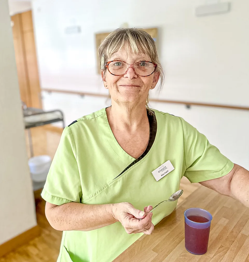Frau mit Brille und grüner Pflegekleidung sitzt lächelnd an einem Holztisch, hält einen Löffel neben einem Becher.