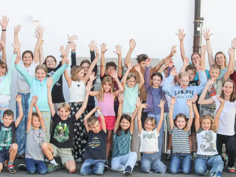 Eine grosse Gruppe von Kindern und Erwachsenen steht jubelnd mit erhobenen Armen vor einer Hauswand.