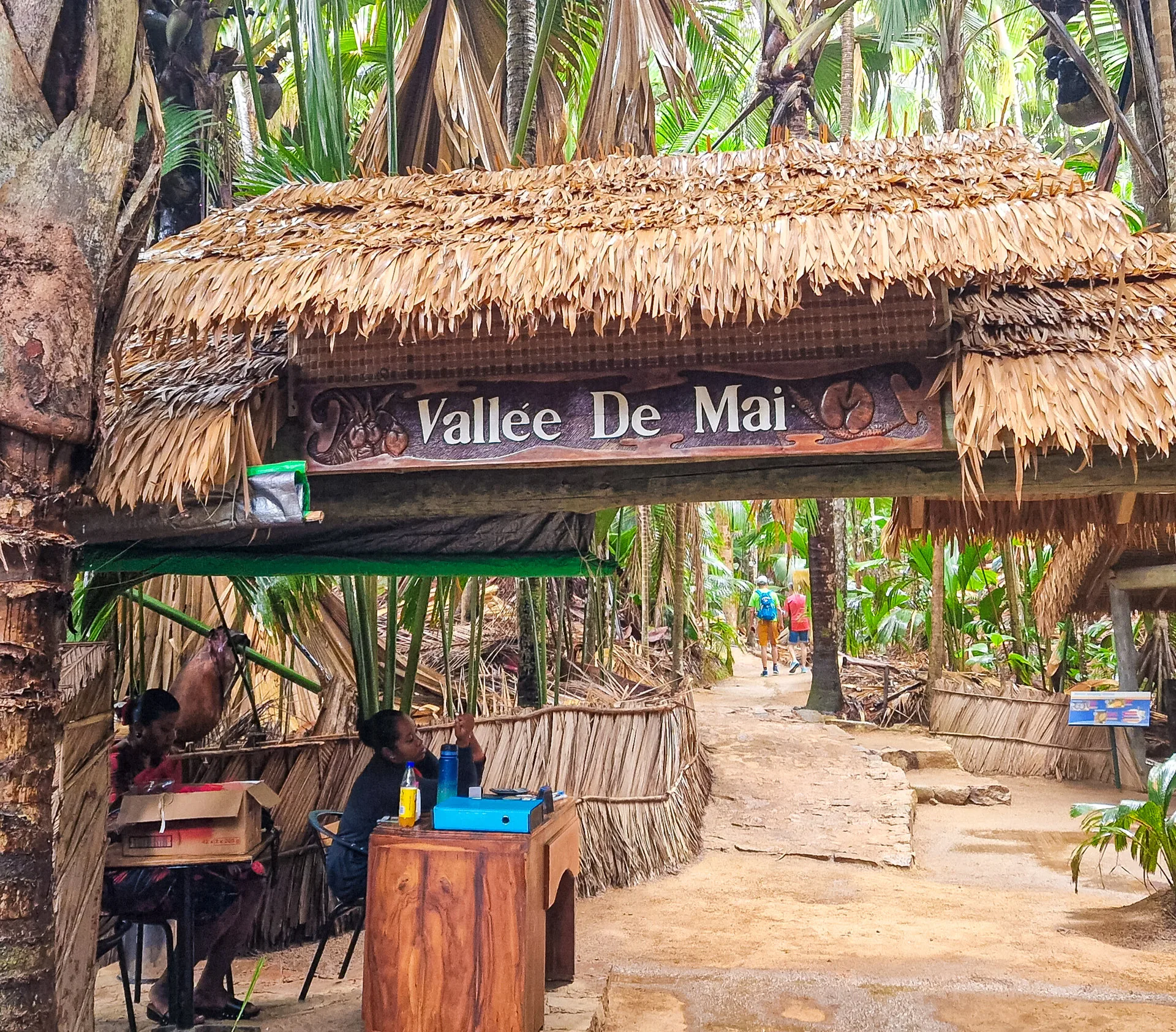 Ein strohgedecktes Torgebäude mit der Aufschrift „Vallée De Mai“, umgeben von tropischem Palmenwald.