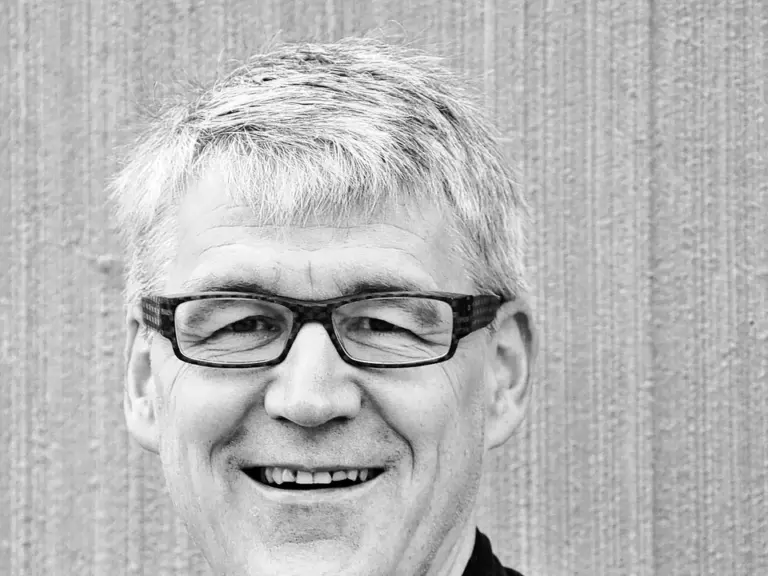 Ein lächelnder Mann mittleren Alters mit Brille und grauem Haar in dunklem Hemd vor gestreifter Wand.
