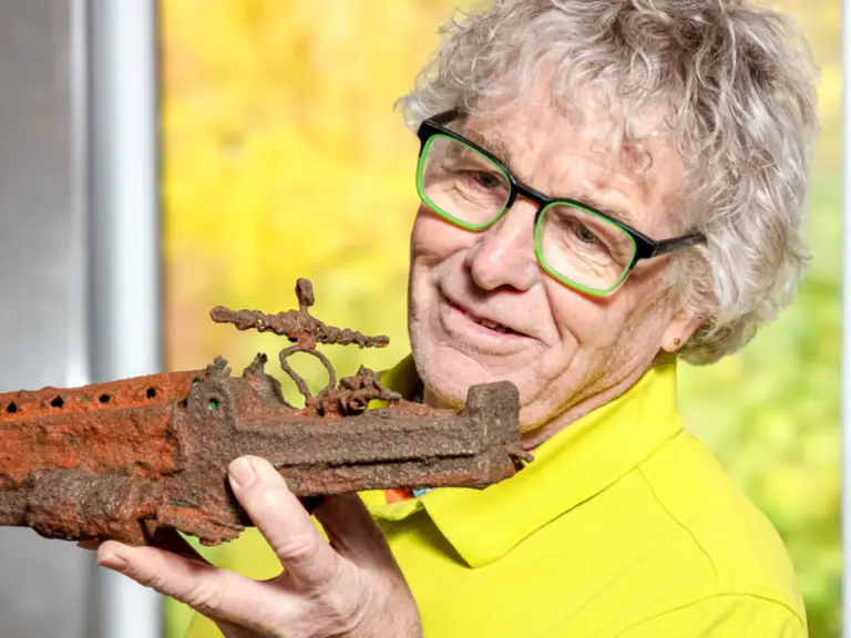Ein lächelnder älterer Mann mit grüner Brille hält ein stark verrostetes altes Metallobjekt in den Händen.