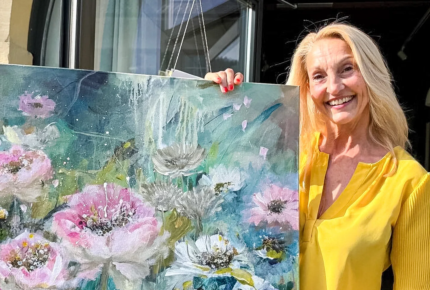 Eine lächelnde Frau mittleren Alters in gelber Bluse steht neben einem gemalten Blumenbild.