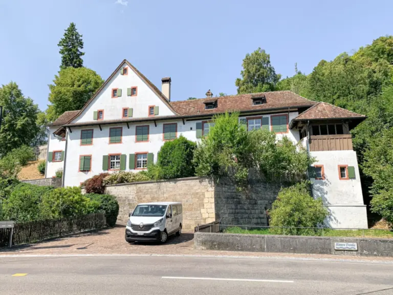 Ein grosses weisses Fachwerkhaus mit rotem Ziegeldach und grünen Fensterläden, umgeben von Bäumen, davor parkt ein Kleinbus.