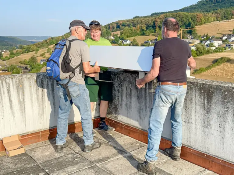 Drei Männer befestigen auf einer Mauer einen weissen Kasten, im Hintergrund hügelige Landschaft.