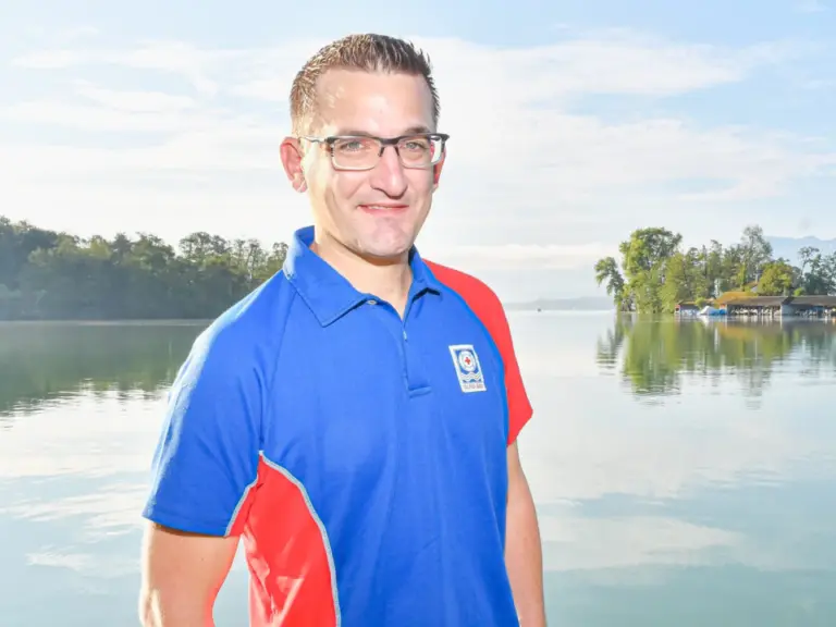 Ein Mann mit Brille und Polo-Shirt lächelt vor einem ruhigen See mit bewaldeten Ufern.