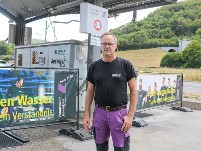 Ein Mann mit Brille und schwarzem T-Shirt steht vor einer Autowaschanlage mit Werbebannern.