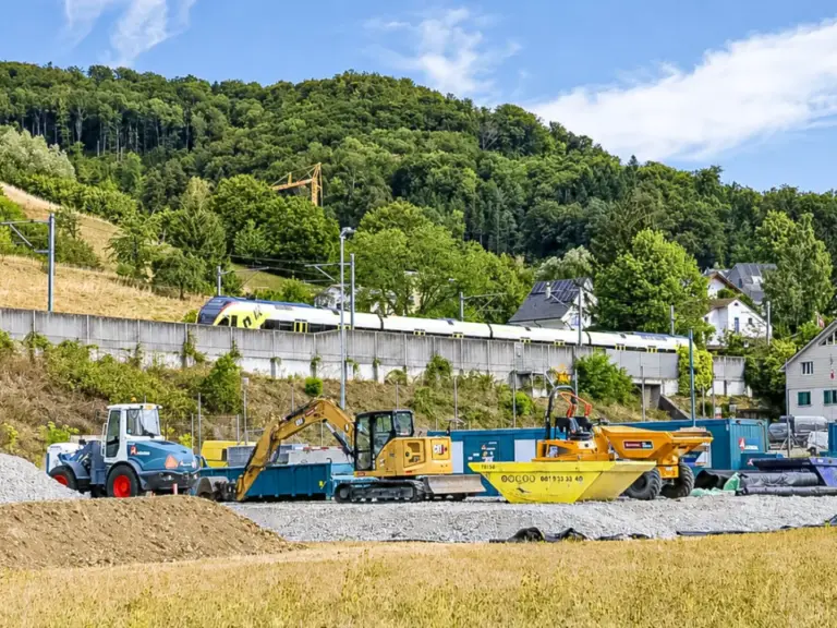 Baustelle mit Bagger und Radlader vor einer Böschung, im Hintergrund fährt ein Zug über eine Brücke.