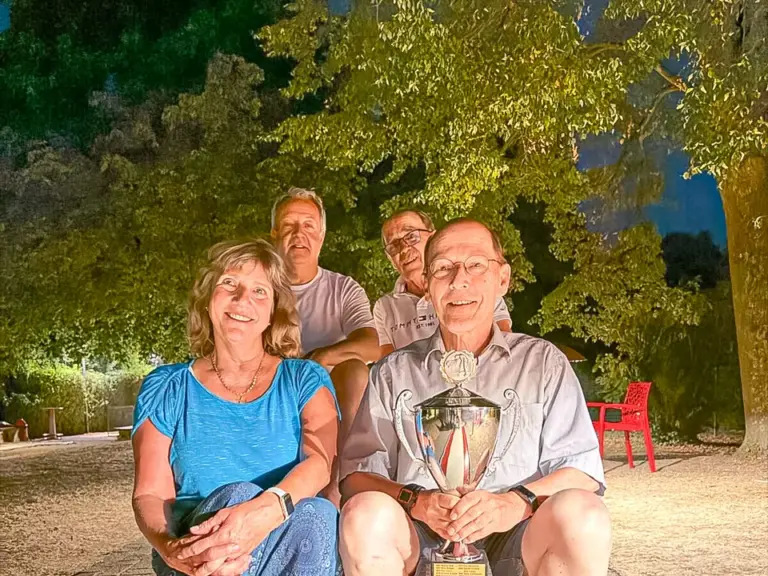 Vier lächelnde Personen sitzen abends im Freien auf einer Bank, der Mann vorne rechts hält einen Pokal.