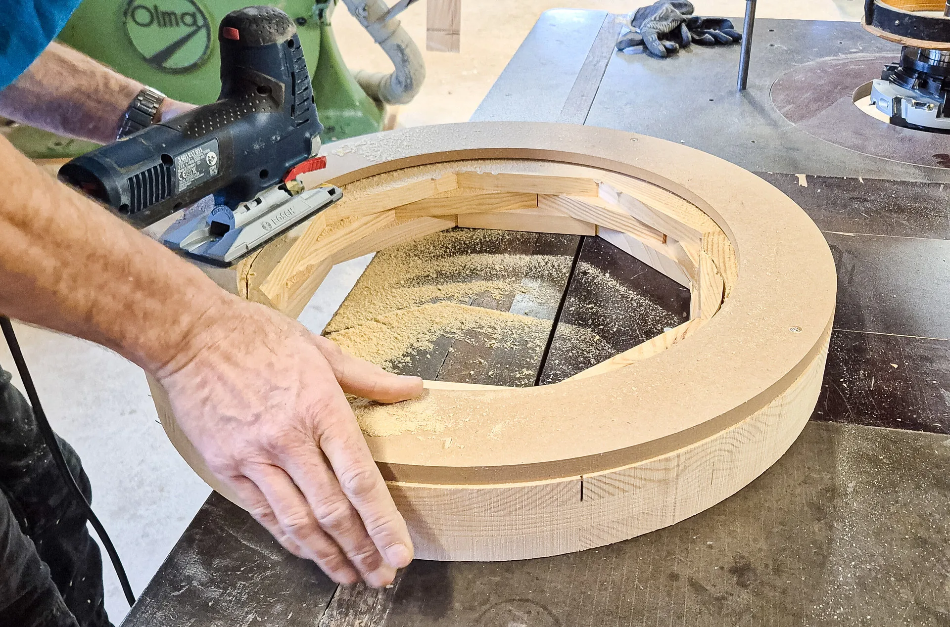 Eine Hand hält einen runden Holzring auf einer Werkbank fest, daneben Sägestaub und Werkzeug.