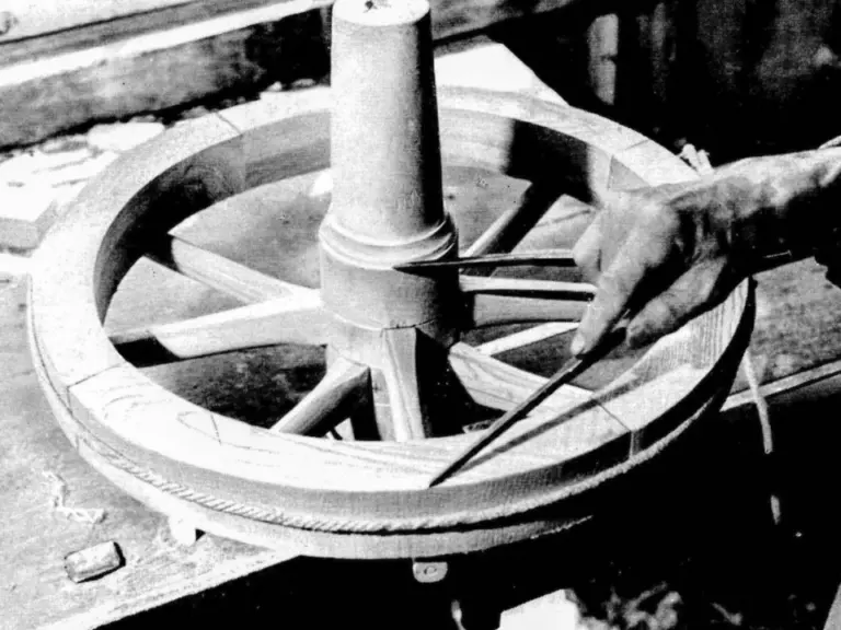 Eine Hand hält einen Zirkel an ein hölzernes Wagenrad mit Speichen und zentraler Nabe.