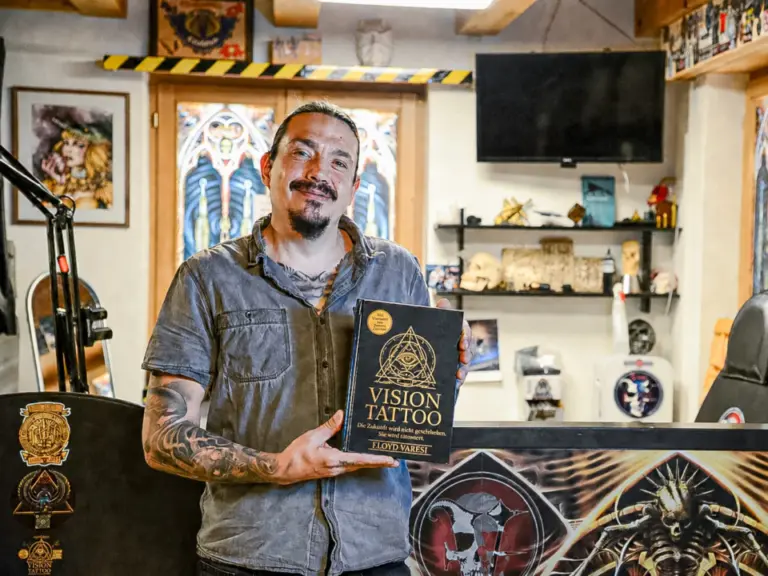 Ein tätowierter Mann mit Bart hält lächelnd ein Buch mit dem Titel «Vision Tattoo» in einem Tattoostudio.