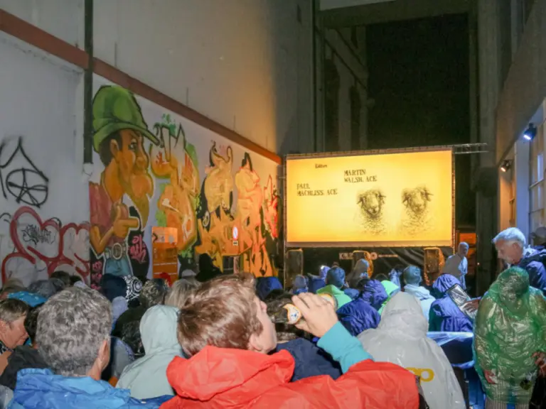 Menschen in Regenjacken sitzen nachts im Freien vor einer beleuchteten Kinoleinwand an einer bemalten Mauer.