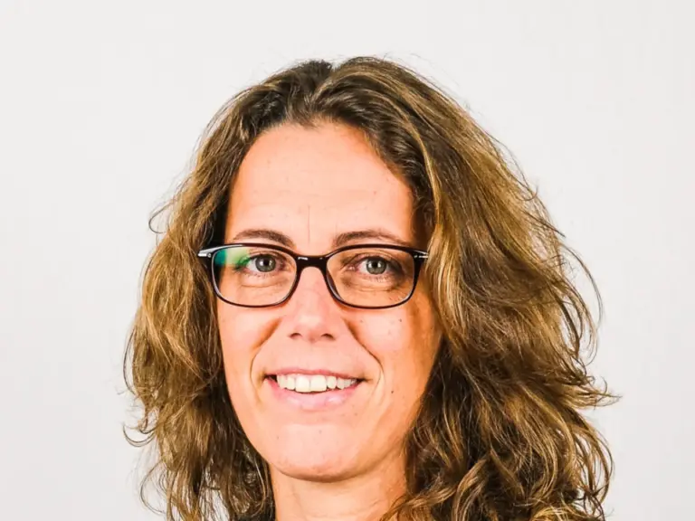Frau mit lockigem, schulterlangem Haar und Brille lächelt in die Kamera, im dunklen Blazer.