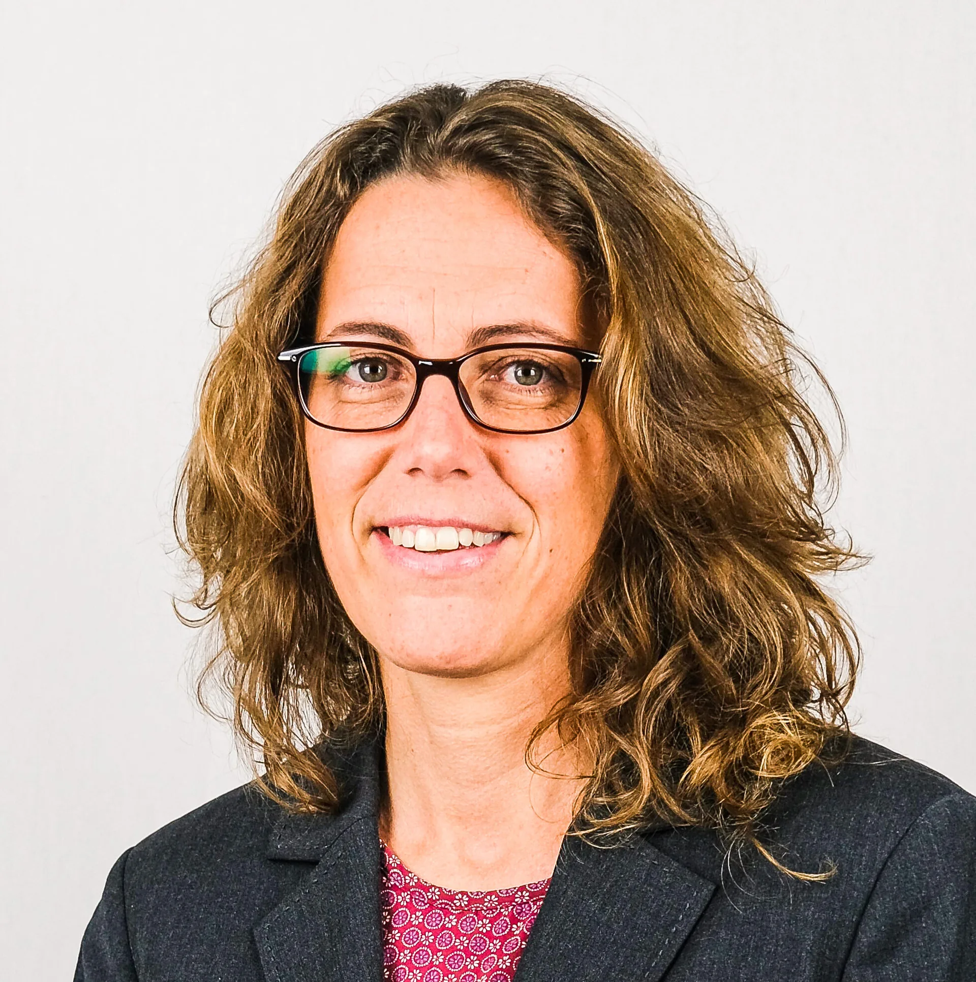 Frau mit lockigem, schulterlangem Haar und Brille lächelt in die Kamera, im dunklen Blazer.