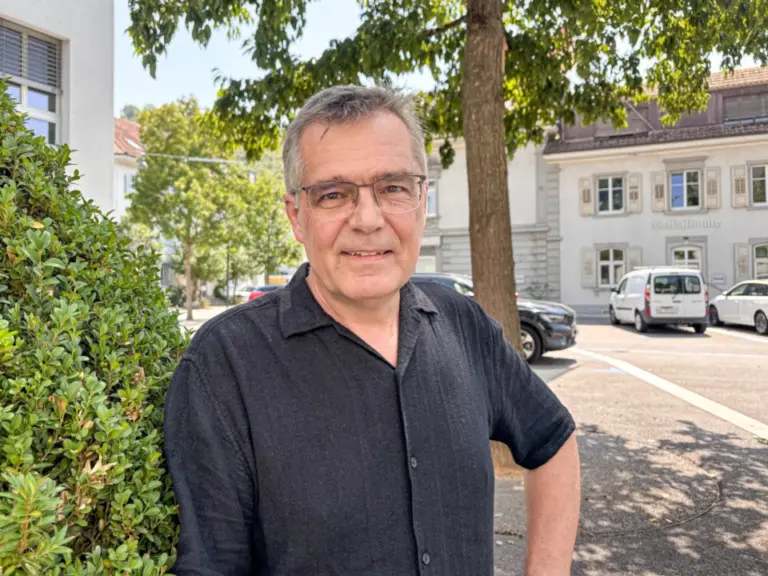Ein Mann mit Brille und grauem Haar in dunklem Hemd steht lächelnd vor einer Hecke auf einem Platz.