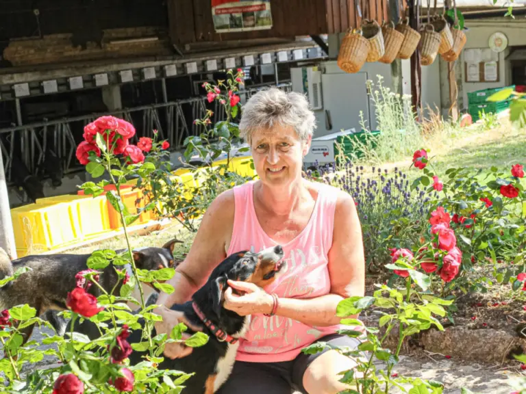 Eine kniende Frau hält einen Hund im Rosenbeet, im Hintergrund ein zweiter Hund unter einer Überdachung.