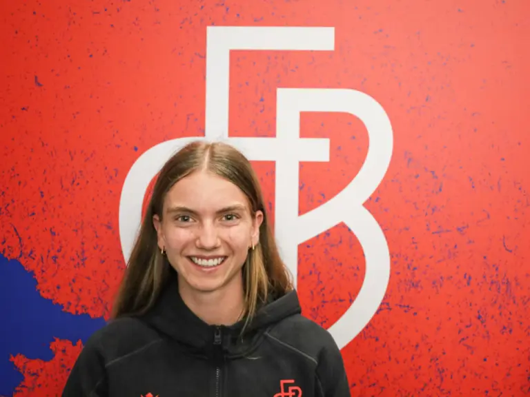Eine lächelnde junge Frau mit langen Haaren steht vor einer roten Wand mit grossem Logo.