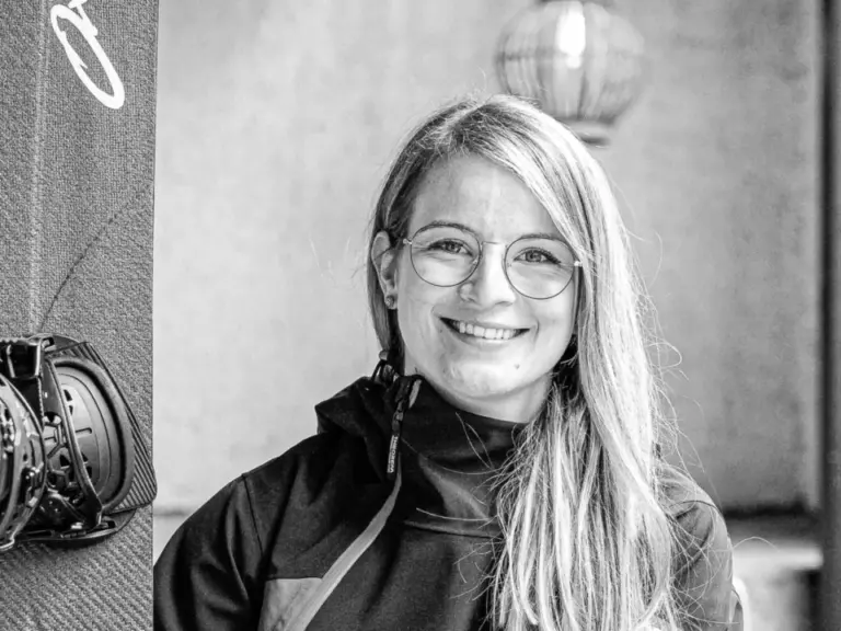 Lächelnde blonde Frau mit Brille sitzt auf einer Bank neben einem Snowboard mit Fernbedienung.