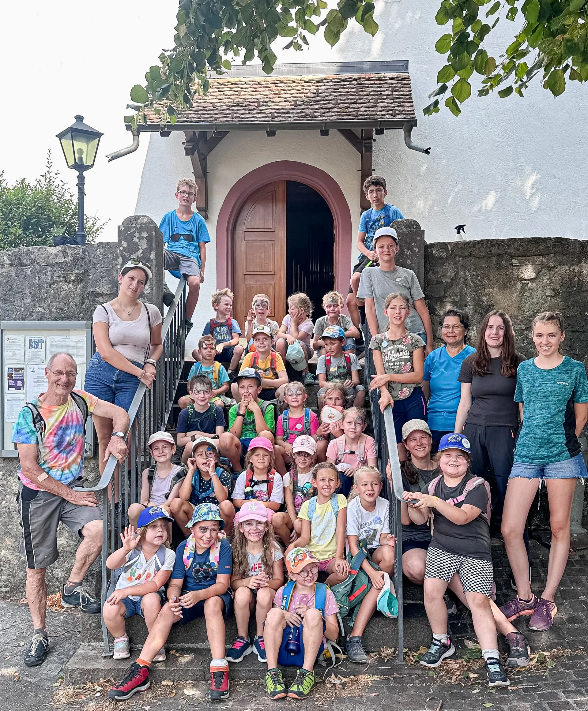 Grosse Gruppe von Kindern und Erwachsenen sitzt und steht auf einer Treppe vor einem Kirchenportal.