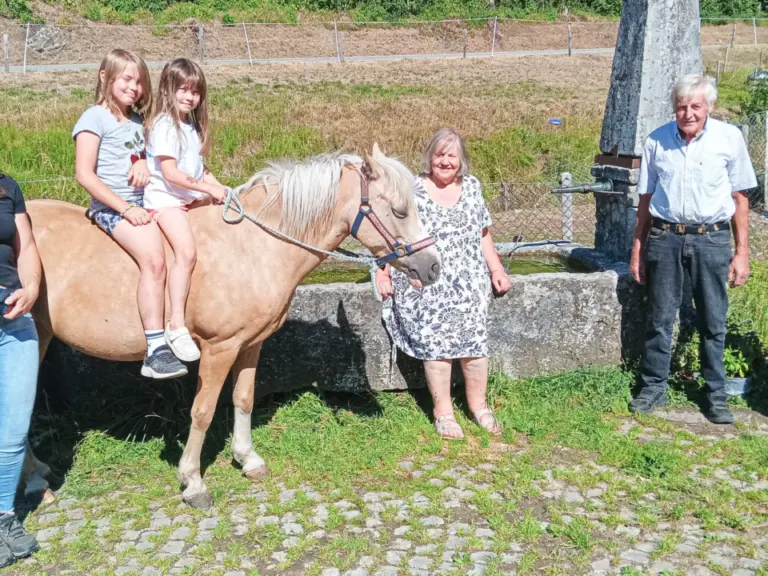 Sechs Personen stehen bei einem Steintrog, zwei Mädchen sitzen auf einem Pony, im Hintergrund Grün und ein steinernes Denkmal.