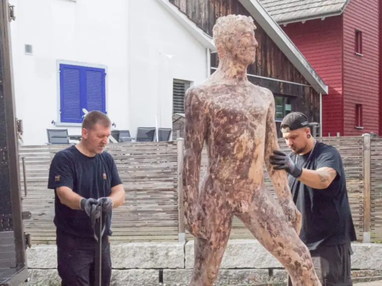 Zwei Männer stellen eine grosse menschenförmige Skulptur aus Metall vor einem Lastwagen auf.