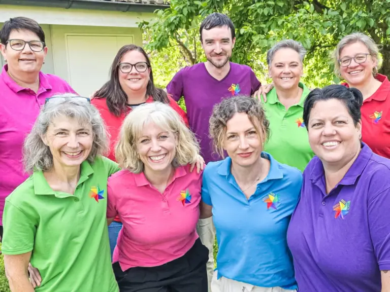 Acht lächelnde Personen in bunten Poloshirts stehen und knien im Garten vor Bäumen.