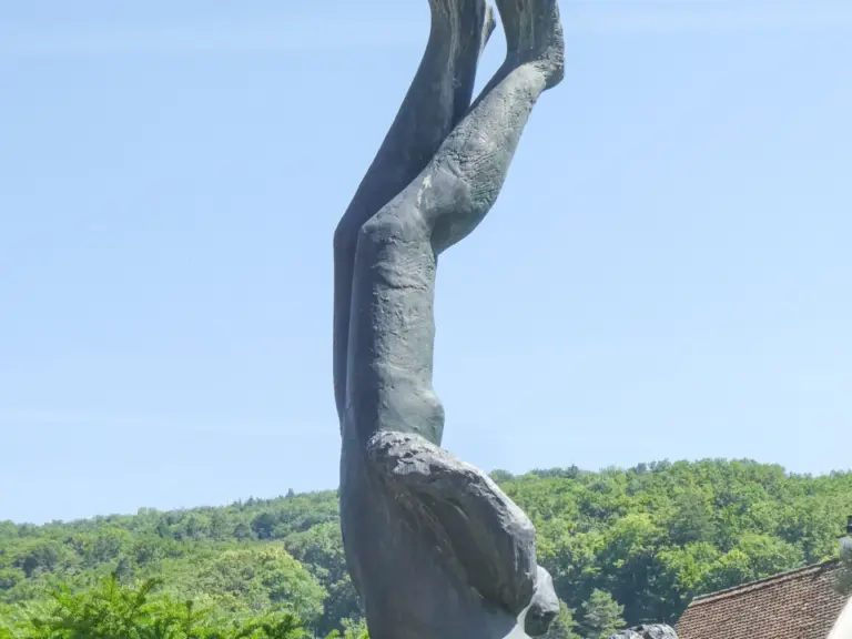 Eine Bronzestatue zeigt einen Menschen kopfüber mit erhobenen Beinen, umgeben von blühenden Sträuchern vor Waldhügeln.