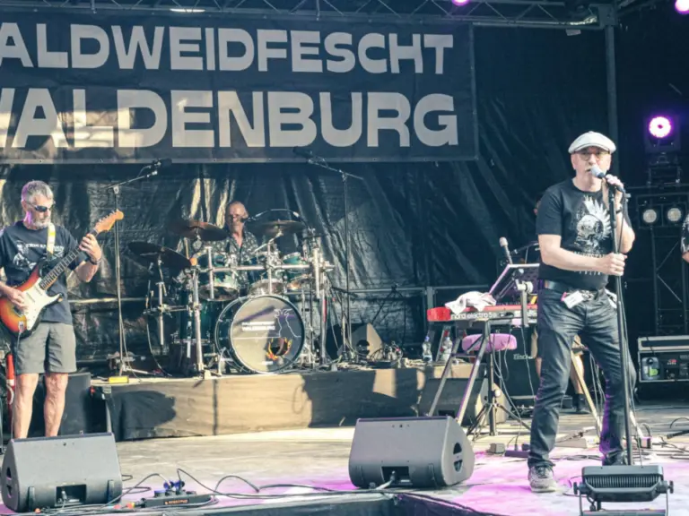 Eine Band spielt live auf einer Bühne mit Gitarren, Schlagzeug und Gesang vor einem Bühnenbanner.
