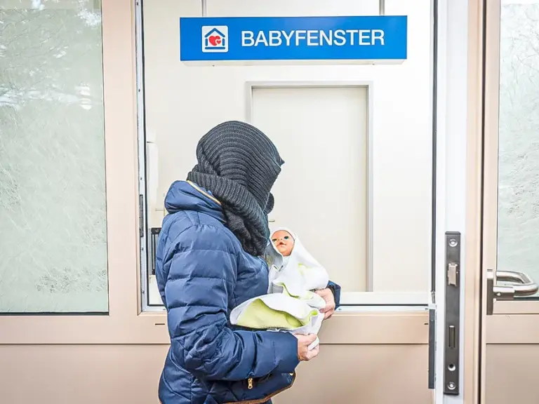Eine vermummte Person hält ein Baby vor einer Tür mit der Aufschrift „Babyfenster“.