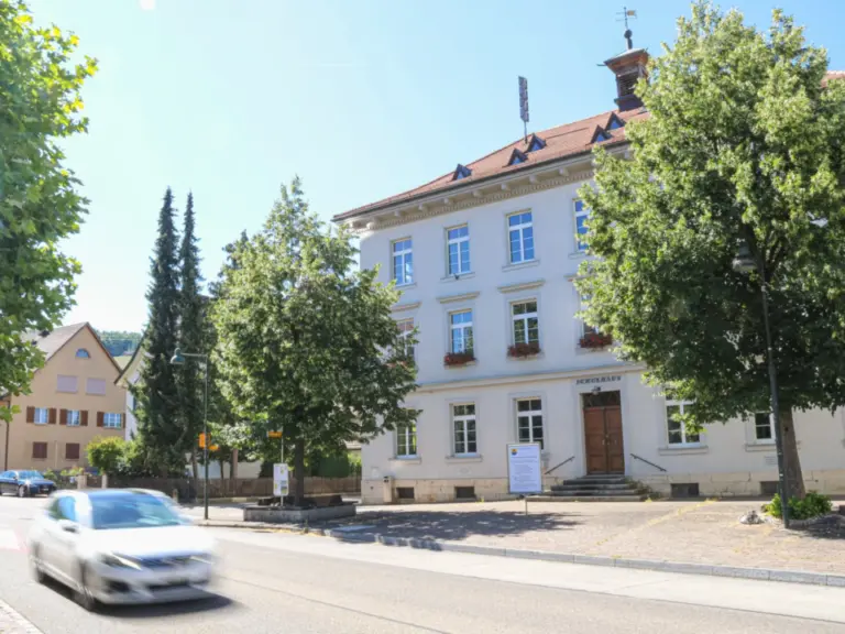 Historisches, weiss verputztes Gebäude mit rotem Ziegeldach hinter Bäumen, davor fährt ein Auto vorbei.