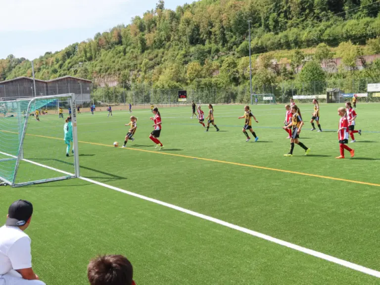 Junge Fussballerinnen kämpfen auf einem Kunstrasenplatz um den Ball, im Hintergrund bewaldete Hügel.