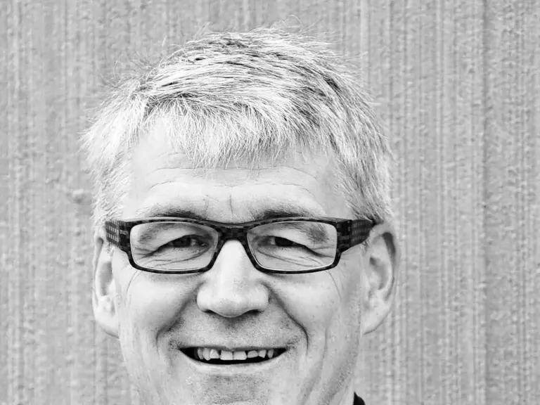 Ein älterer Mann mit grauem Haar und Brille lächelt, er trägt ein dunkles Hemd.