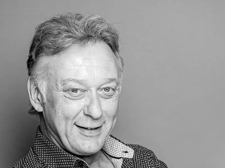 Ein lächelnder älterer Mann mit verschränkten Armen in gemustertem Hemd, Schwarzweißaufnahme.