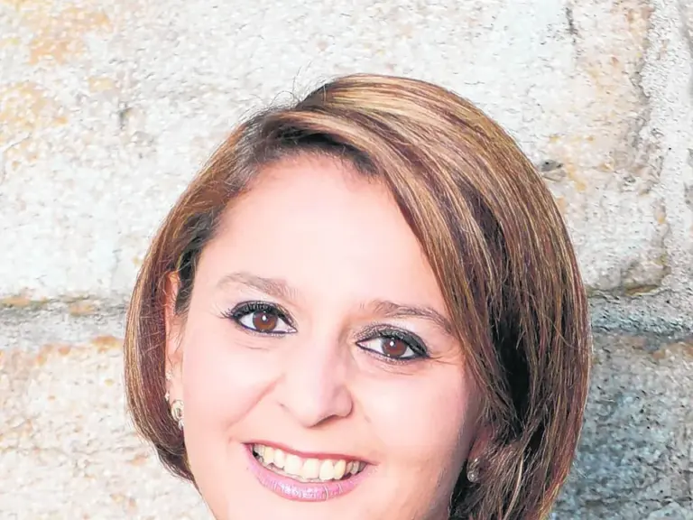 Eine lächelnde Frau mit schulterlangem braunem Haar trägt einen schwarzen Blazer vor einer Steinwand.