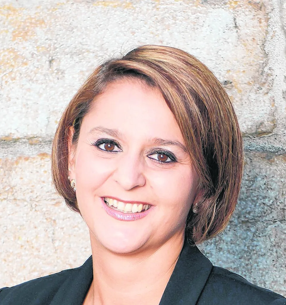 Eine lächelnde Frau mit schulterlangem braunem Haar trägt einen schwarzen Blazer vor einer Steinwand.