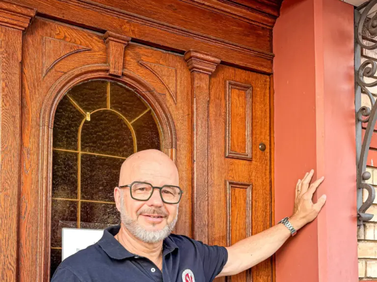 Ein lächelnder Mann mit Brille und Glatze steht vor einer alten Holztür und lehnt eine Hand daran.