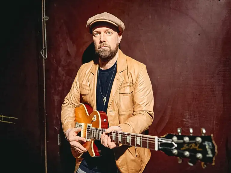 Ein Mann mit Schiebermütze und Bart hält eine E-Gitarre und blickt in die Kamera.