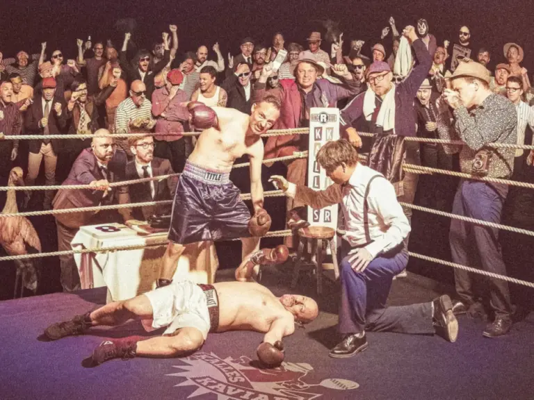 Ein Boxer steht triumphierend über einem am Boden liegenden Gegner im Ring, jubelnde Zuschauer im Hintergrund.