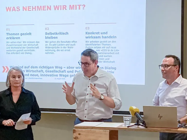 Drei Personen stehen an einem Rednerpult vor einer Leinwand mit Präsentationsfolie im Vortragssaal.