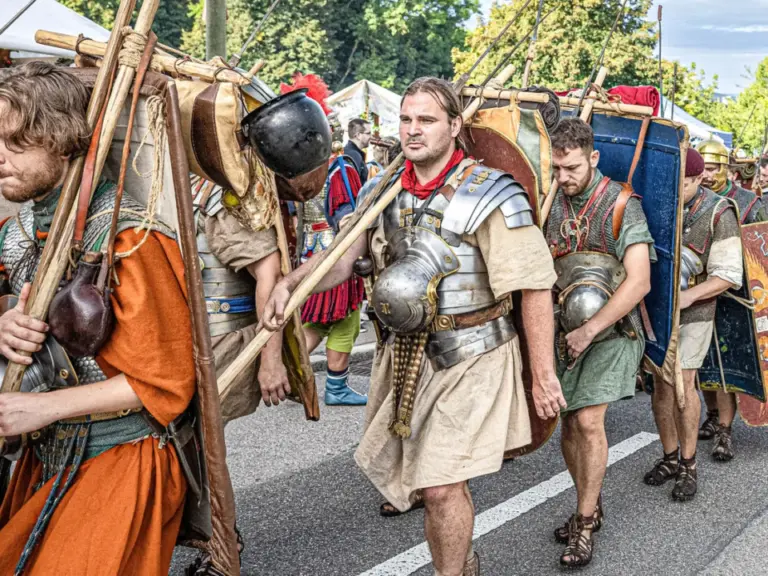 Männer in römischen Legionärskostümen mit Schilden und Speeren marschieren in einer Prozession durch eine Straße.