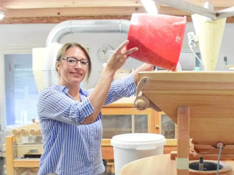 Eine Frau in gestreifter Bluse giesst Getreide aus einem roten Eimer in eine hölzerne Steinmühle.