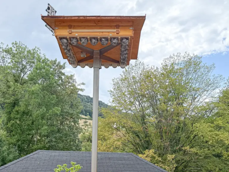 Fünf Personen in rot-gelber Kleidung stehen nebeneinander vor einem Pavillon unter einem hohen Holzmast mit Dach.