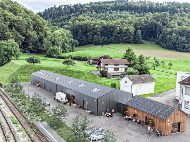 Luftaufnahme einer langen Lagerhalle mit Solaranlage auf dem Dach, umgeben von Wiesen und Wald.