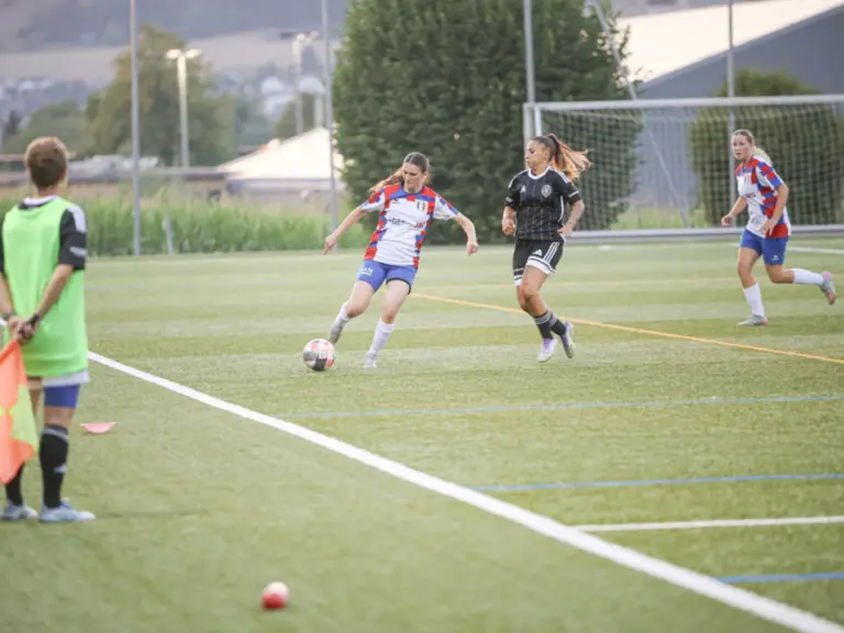 Fussballerinnen laufen einem Ball auf dem Rasen hinterher, im Vordergrund steht ein Linienrichter mit Fahne.