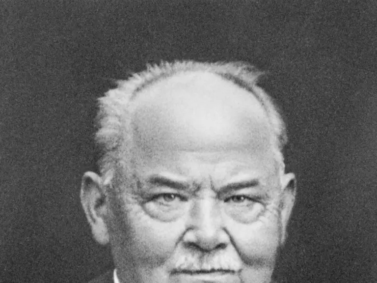 Schwarz-weiß-Porträt eines älteren Mannes mit Halbglatze, Schnurrbart, Anzug und Krawatte, ernster Blick in die Kamera.