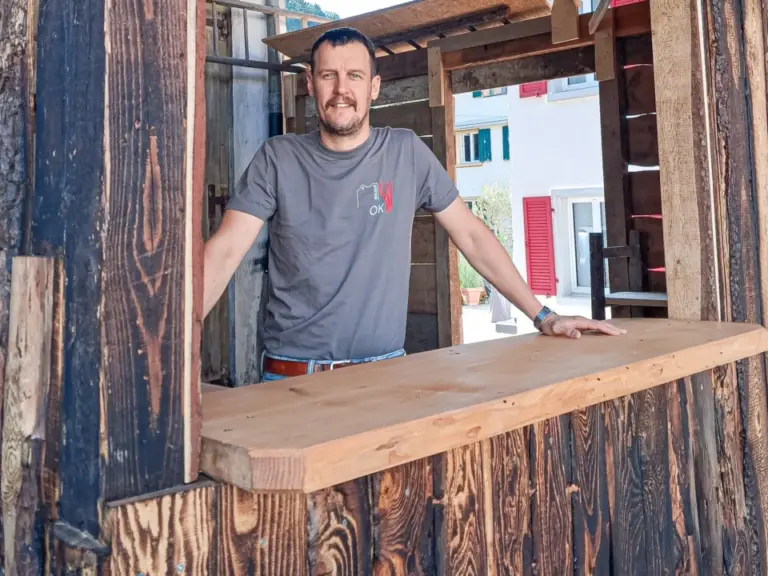 Ein Mann mit Bart lehnt an einem Holztresen in einer rustikalen Hütte aus Holzbalken.