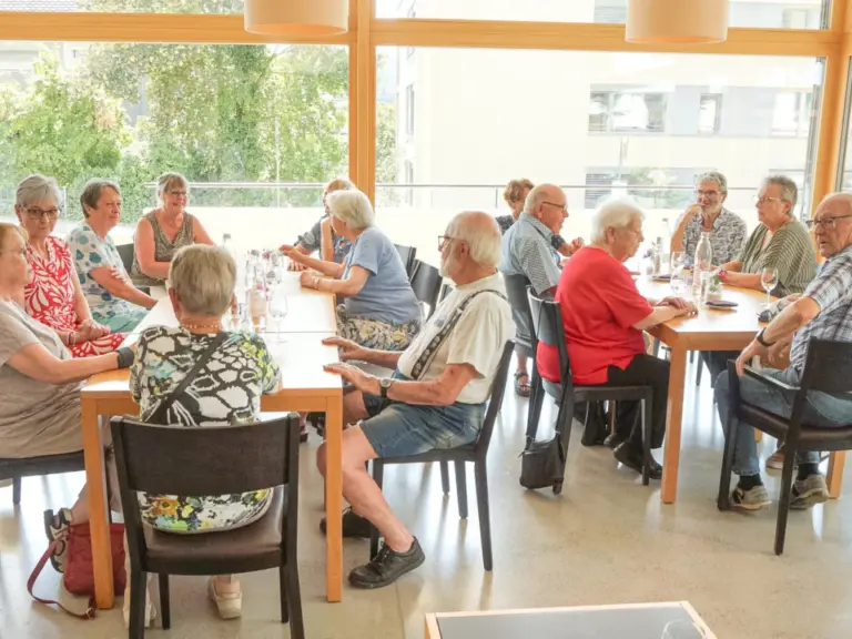 Eine Gruppe älterer Menschen sitzt gemeinsam an Tischen in einem hellen Raum mit grossen Fenstern.