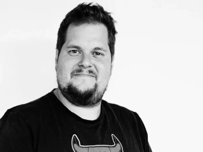 Schwarz-weiss-Porträt eines lächelnden Mannes mit Bart und dunklem T-Shirt vor heller Wand.