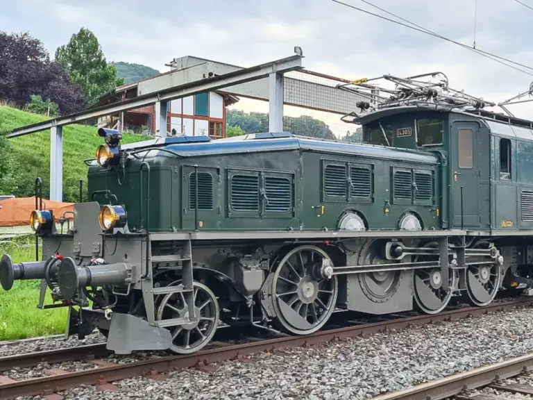 Eine dunkelgrüne historische Elektrolokomotive mit markanten Rädern steht auf Gleisen unter Fahrleitungen.